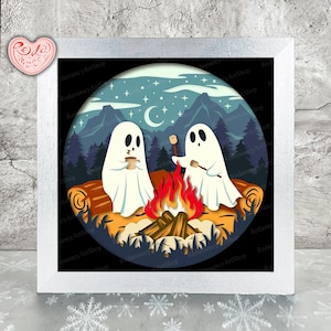 Op de afbeelding: Een ingelijste kunstdruk met twee cartoongeesten die marshmallows roosteren boven een kampvuur. De scène speelt zich af in een bos met bergen, een halve maan en sterren. Het kunstwerk is in een cirkelvormig ontwerp met een witte lijst.