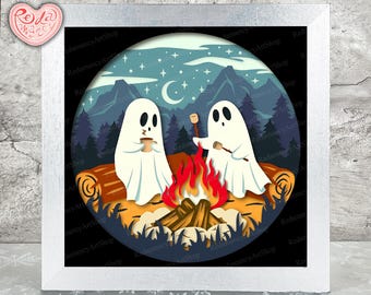 Op de afbeelding: Een ingelijste kunstdruk met twee cartoongeesten die marshmallows roosteren boven een kampvuur. De scène speelt zich af in een bos met bergen, een halve maan en sterren. Het kunstwerk is in een cirkelvormig ontwerp met een witte lijst.