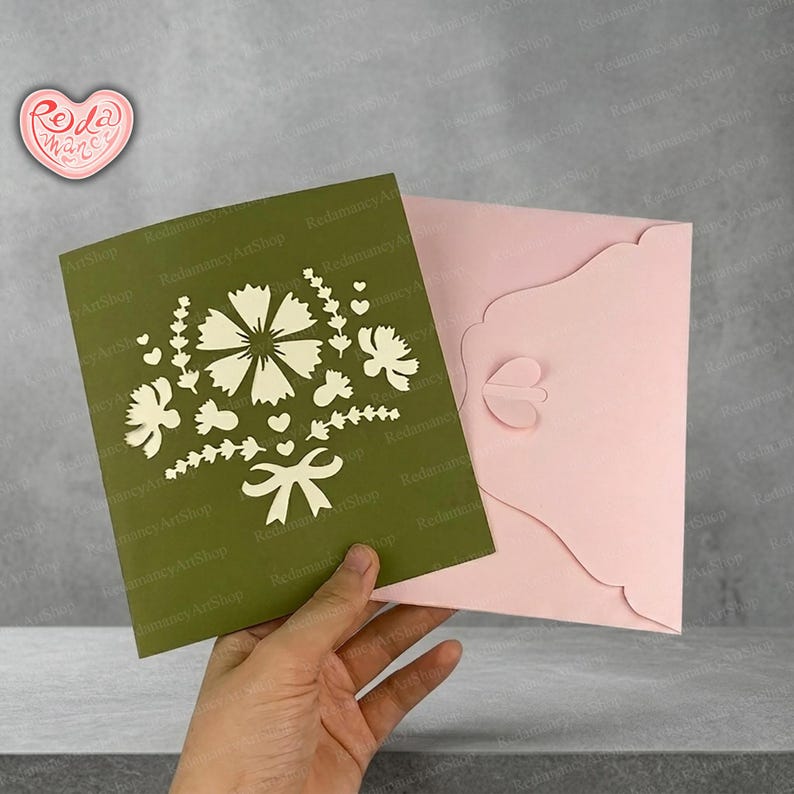 May include: A handmade pop-up greeting card with a floral bouquet design. The card is olive green with a white cut-out floral design and a pink envelope. The card is from Redamancy ArtShop.
