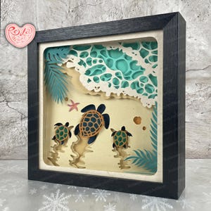 May include: A framed 3D paper art piece depicting a beach scene with three sea turtles. The artwork features layered paper cutouts showing waves, sand, palm leaves, and a starfish. The frame is black.