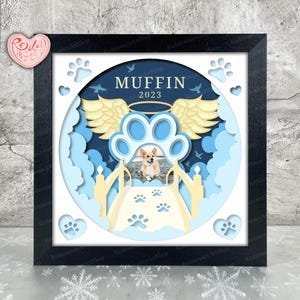 Könnte beinhalten: Gerahmtes Gedenkkunstwerk mit einem Corgi-Hund. Das Design zeigt einen Pfotenabdruck mit Engelsflügeln, Wolken und dem Namen "MUFFIN" mit dem Jahr "2023". Das Kunstwerk ist aus blauen und weißen Papierschnitten gefertigt und in einem schwarzen Rahmen gefasst.