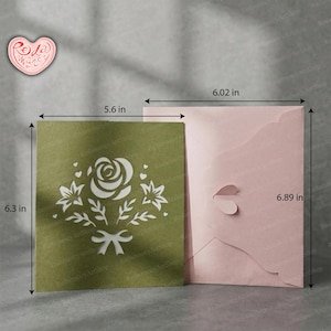 May include: A green card (14.2 x 16 cm) with a white floral design and a pink envelope (15.3 x 17.5 cm). The card features a cut-out rose, leaves, and a bow. The envelope has a heart-shaped closure.