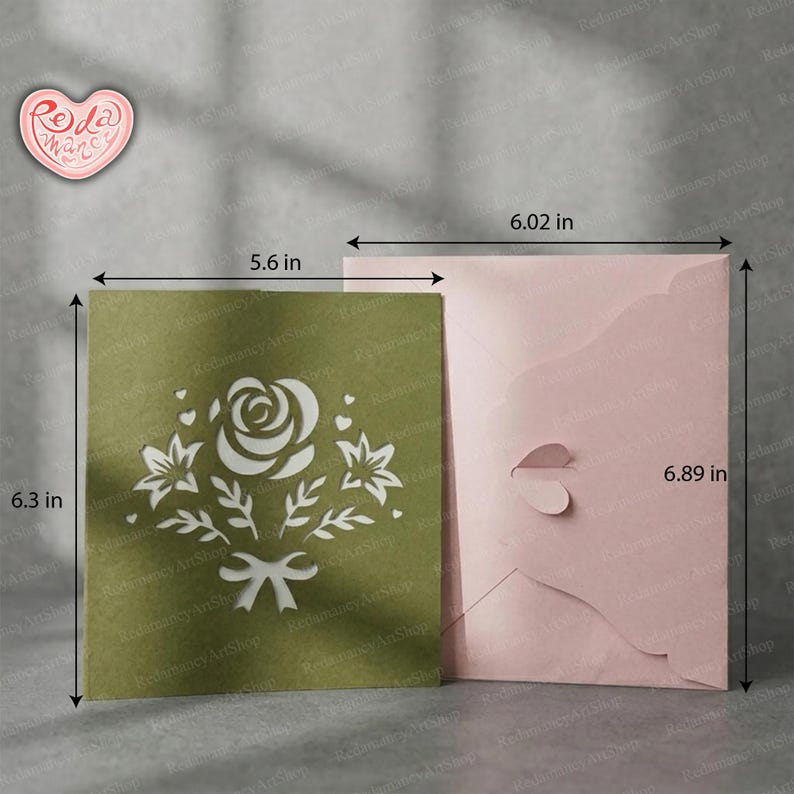 May include: A green card (14.2 x 16 cm) with a white floral design and a pink envelope (15.3 x 17.5 cm). The card features a cut-out rose, leaves, and a bow. The envelope has a heart-shaped closure.