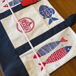 May include: A cream-colored canvas tote bag with dark navy blue straps and trim, featuring red and blue fish illustrations and a red circular Trader Joe's logo. A white measuring tape shows the bag length is approximately 22 inches long.