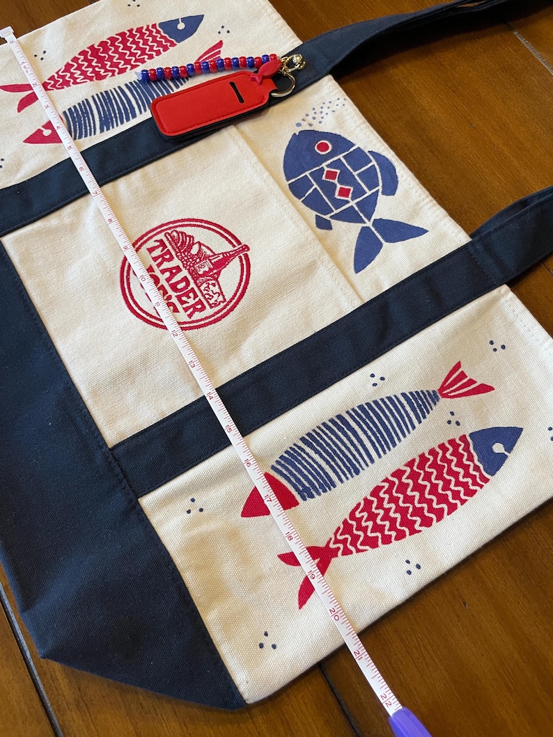 May include: A cream-colored canvas tote bag with dark navy blue straps and trim, featuring red and blue fish illustrations and a red circular Trader Joe's logo. A white measuring tape shows the bag length is approximately 22 inches long.