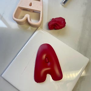 May include: A light pink plastic letter A cookie cutter, a small ball of red clay, a silver metal craft tool, and a red clay letter A shape. The red clay letter A is pressed into a smooth, rounded font style with a small center cutout.