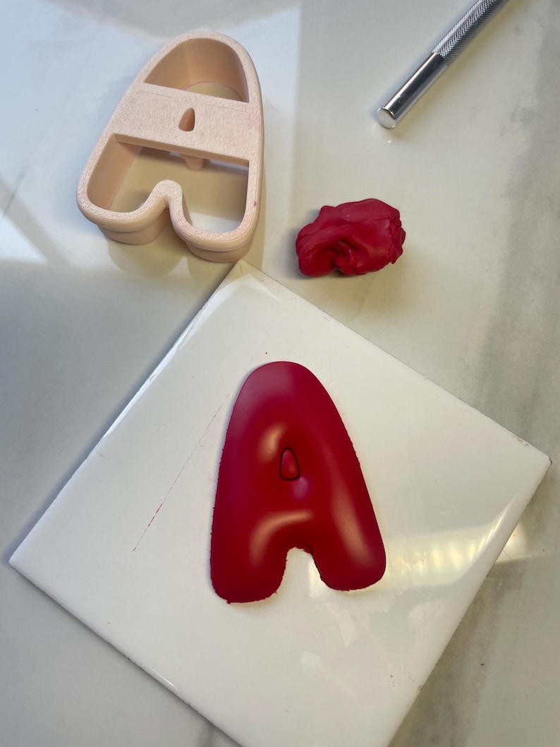 May include: A light pink plastic letter A cookie cutter, a small ball of red clay, a silver metal craft tool, and a red clay letter A shape. The red clay letter A is pressed into a smooth, rounded font style with a small center cutout.