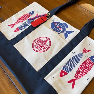 May include: A cream-colored canvas tote bag with navy blue trim and handles. The bag features red and blue fish illustrations, a circular Trader Joe's logo in red, and a red rectangular luggage tag attached with a blue and red beaded chain.
