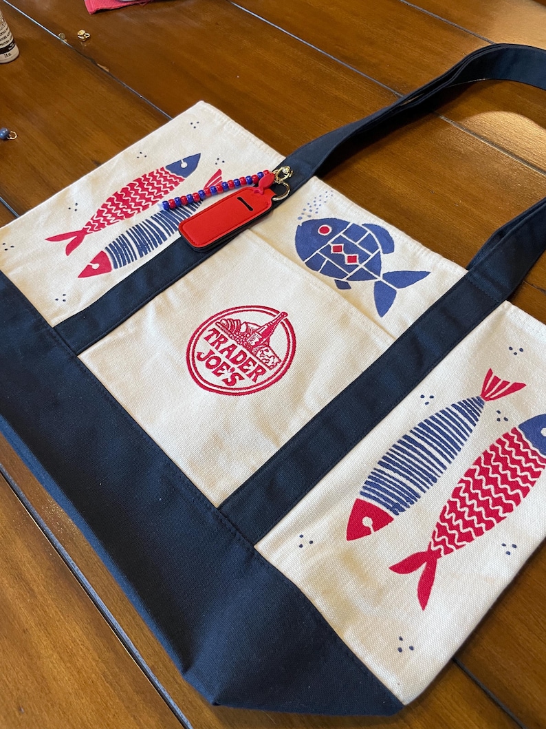 May include: A cream-colored canvas tote bag with navy blue trim and handles. The bag features red and blue fish illustrations, a circular Trader Joe's logo in red, and a red rectangular luggage tag attached with a blue and red beaded chain.
