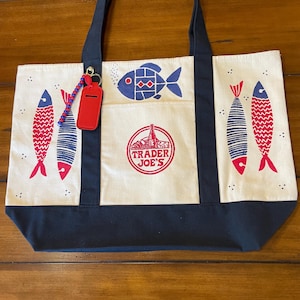 May include: A cream-colored canvas tote bag with dark navy blue handles and a bottom panel. The bag features a red and navy blue fish print, a central circular Trader Joe's logo, and a red luggage tag attached with a blue and red beaded keychain.