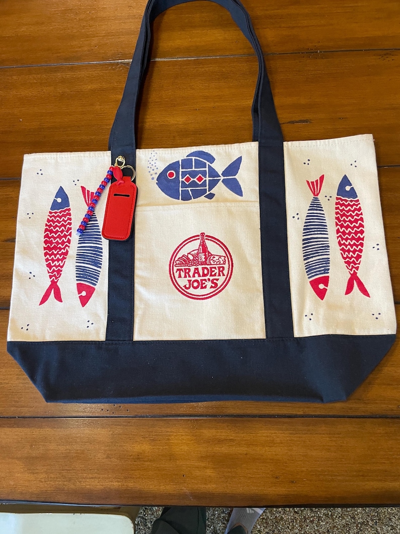 May include: A cream-colored canvas tote bag with dark navy blue handles and a bottom panel. The bag features a red and navy blue fish print, a central circular Trader Joe's logo, and a red luggage tag attached with a blue and red beaded keychain.