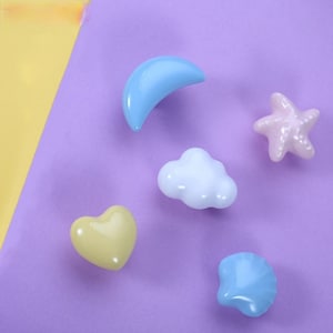 May include: Five pastel-colored ceramic drawer pulls in the shapes of a crescent moon, a cloud, a heart, a seashell, and a starfish. The drawer pulls are on a purple background.