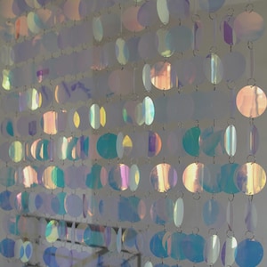 Op de afbeelding: Een glinsterend gordijn van iriserende, ronde pailletten hangt verticaal. De pailletten zijn verbonden door kleine metalen ringen, waardoor een vloeiend, decoratief element ontstaat. De kleuren verschuiven van lichtroze en geel naar blauw en groen, waardoor het licht wordt gereflecteerd.