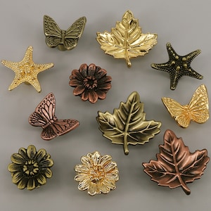 Op de afbeelding: Assortiment van decoratieve kastknoppen in verschillende vormen en kleuren, waaronder vlinders, bladeren en bloemen. De knoppen zijn van metaal en hebben een vintage uitstraling.