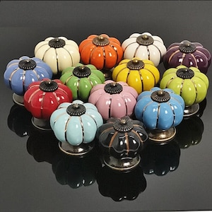 Pode incluir: Conjunto de 14 botões de gaveta de cerâmica em forma de abóbora coloridos com detalhes dourados. Os botões estão dispostos em círculo sobre uma superfície preta.