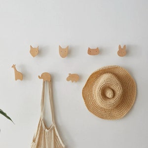 Peut inclure: Crochets muraux en bois en forme de girafe, d'éléphant, d'hippopotame, de renard, de chat et de lapin. Les crochets sont fixés à un mur blanc avec un chapeau de paille accroché à l'un des crochets.