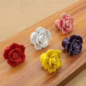 May include: Five ceramic rose-shaped drawer pulls in different colors: red, white, pink, yellow, and purple. The pulls are arranged on a wooden surface.