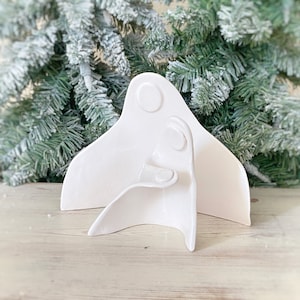 May include: A white ceramic nativity scene with two figures. The larger figure has a rounded top with a circular detail, and the smaller figure is in front. The background features a frosted green Christmas tree.