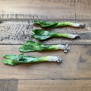 May include: Four ceramic leeks, painted in shades of green and white, are arranged in a row on a wooden surface. The leeks are realistic in appearance and have a slightly textured finish.