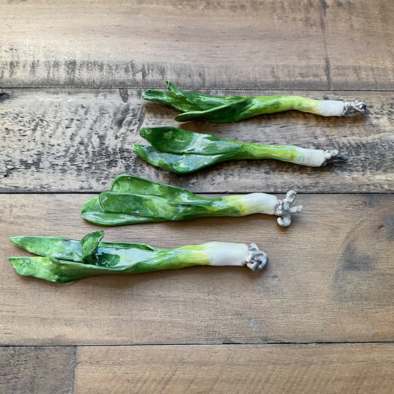 May include: Four ceramic leeks, painted in shades of green and white, are arranged in a row on a wooden surface. The leeks are realistic in appearance and have a slightly textured finish.