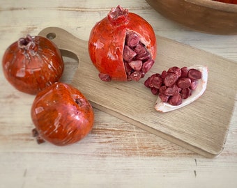 May include: Three ceramic pomegranate sculptures with a glossy red finish. One pomegranate is split open to reveal dark red seeds, with a cluster of seeds resting separately on a wooden board. The other two pomegranates are whole.