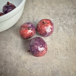 May include: Three round, glossy fruits with a purple and red color scheme. The fruits are arranged in a triangular shape on a gray surface.