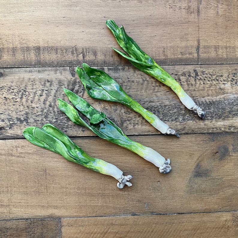 May include: Four ceramic green onions, each with a white bulb at the base, are arranged on a wooden surface.