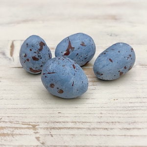 May include: Four blue speckled decorative eggs. Each egg has a smooth, oval shape and is painted in a muted blue color with dark red-brown speckles. The eggs are arranged on a light-colored, weathered wooden surface.
