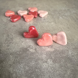 May include: A collection of heart-shaped objects in varying shades of red and pink. The glossy, ceramic-like hearts are scattered across a gray surface, with a few clustered together and others spread out.