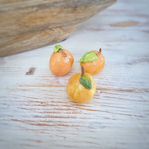 May include: Three glazed, orange-colored marzipan peaches with green leaves and brown stems are arranged on a white-washed wooden surface. A wooden bowl is in the background. The peaches are a dessert or confectionery item.