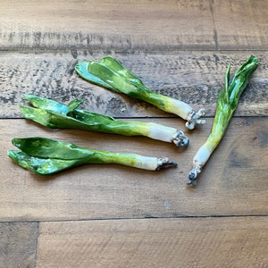 May include: Four ceramic green onions with white bases, arranged on a wooden surface.
