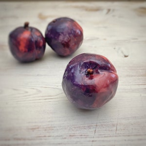 May include: Three plums with a glossy finish, showcasing a blend of deep purple and red hues. The fruits are arranged on a light-colored wooden surface, with one plum in sharp focus and two others slightly blurred in the background.
