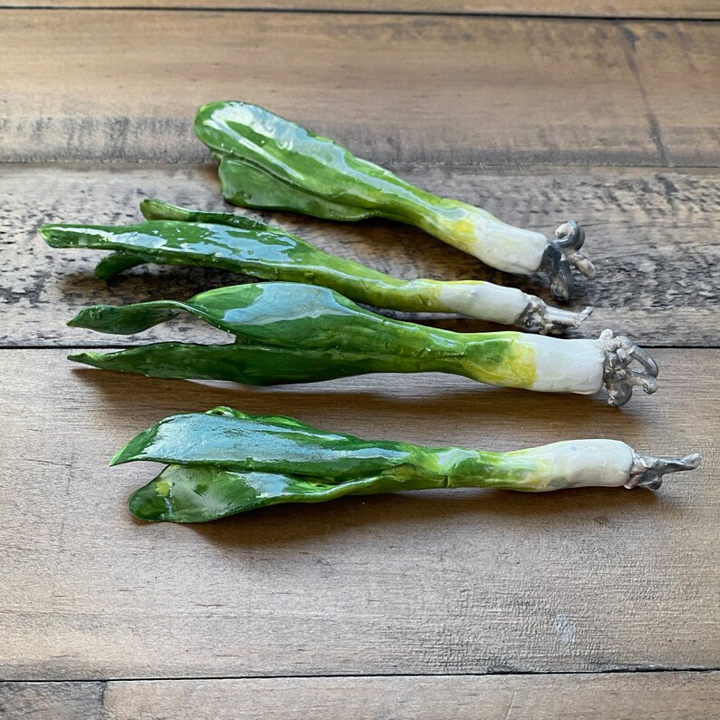 May include: Three ceramic green onions with white bulbs, arranged on a wooden surface.