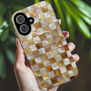 A hand holds the Trendy Gold Mosaic Phone Case by Golden Value SG featuring iridescent gold, white, and beige mosaic tiles with a shimmering, metallic mother-of-pearl effect. Perfect for elegant and unique style. Green foliage is softly blurred in the background.