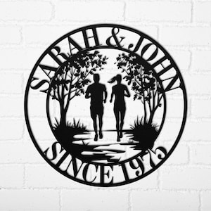 May include: Black metal wall art featuring two runners in a park setting, framed by trees and foliage. The names "SARAH & JOHN" and the text "SINCE 1973" are cut out in a circular design, perfect for a wedding or anniversary gift.