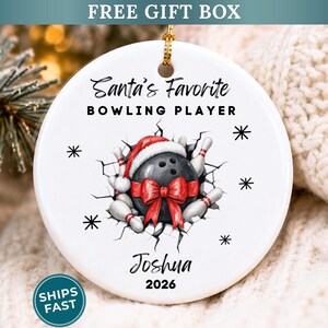 May include: A round white ceramic ornament featuring a black bowling ball wearing a red Santa hat with a red bow, surrounded by white bowling pins. The text reads Santa's Favorite BOWLING PLAYER, Joshua, and 2026, with black snowflake accents.