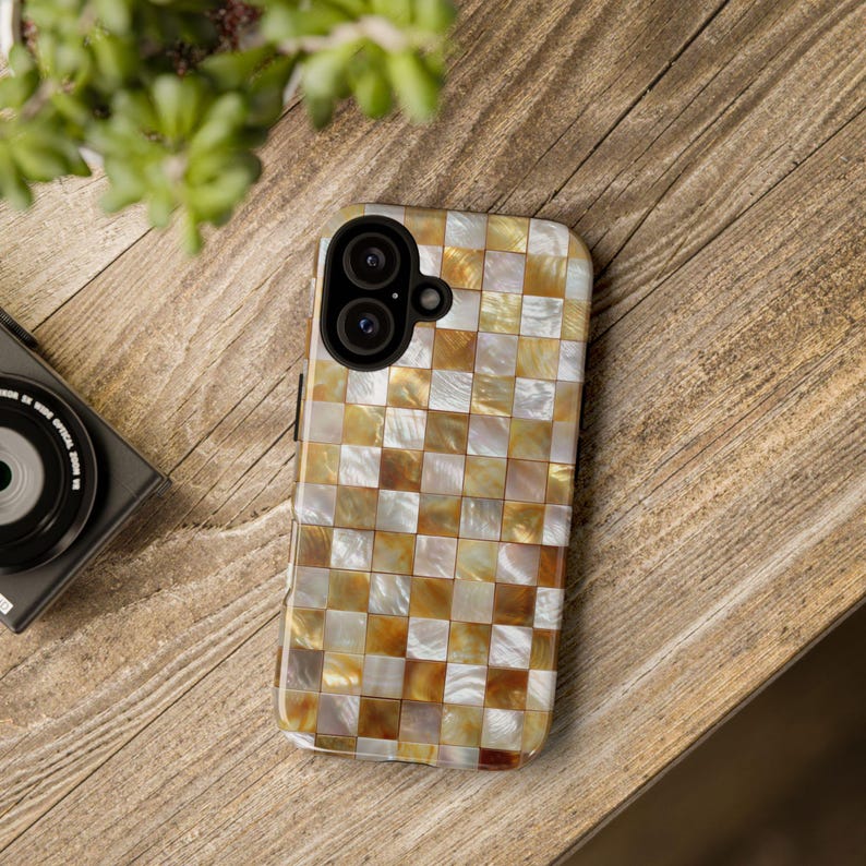 A phone case with a gold and white checkered mosaic pattern rests on a wooden surface next to a camera lens and green plant.