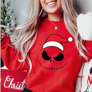 May include: Red Christmas sweater with a black and white Jack Skellington face wearing a Santa hat. The sweater has long sleeves.