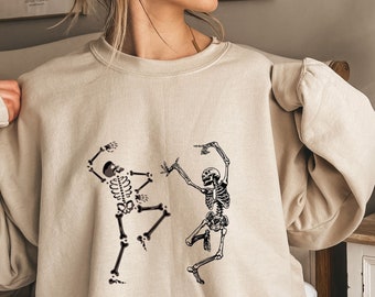 Könnte beinhalten: Beiges Sweatshirt mit einem schwarzen Aufdruck von zwei tanzenden Skeletten. Die Skelette befinden sich in einer lebhaften Pose, eines zeigt und das andere hat die Arme erhoben. Das Sweatshirt hat eine lockere Passform und lange Ärmel.
