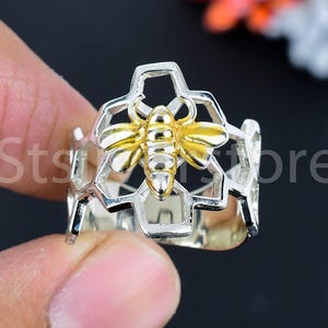 May include: A silver-tone ring featuring a gold-colored bee in the center of a honeycomb design. The ring is held by a hand against a dark background. The text "Sts ore" is visible on the ring.
