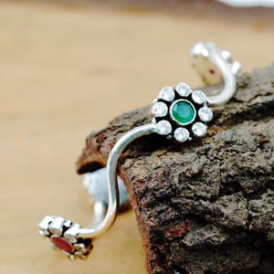 May include: A silver-toned bangle bracelet with a floral design. The center flower features a green gemstone surrounded by clear crystals. The ends of the bracelet have red and clear gemstones. The bracelet is resting on a piece of wood.