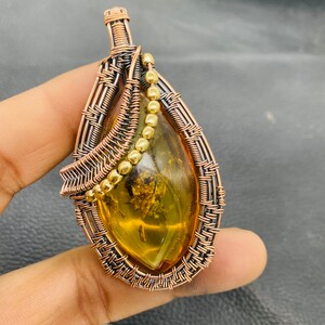 Puede incluir: Colgante artesanal con una piedra de ámbar en forma de lágrima con inclusiones, encapsulada en alambre de cobre. El trabajo de alambre tiene un diseño tejido con detalles de cuentas doradas. El colgante tiene una argolla para colgar.