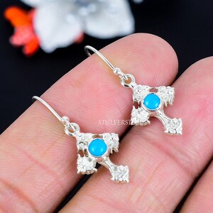 May include: A pair of silver-colored cross earrings with a turquoise blue stone in the center of each cross. The earrings have a decorative, ornate design. The earrings are shown against a blurred background.