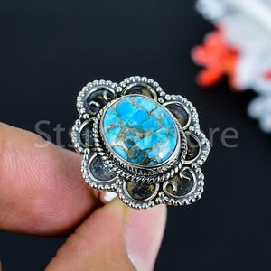 May include: A silver ring featuring an oval turquoise stone with gold flecks, set in a decorative, flower-shaped frame. The ring has a detailed, ornate design with dark accents and a textured band. The word "Store" is partially visible.