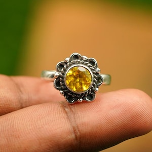 May include: A silver ring with a large, round, yellow gemstone at the center. The gemstone is surrounded by a decorative, flower-like setting with a textured, antique finish. The ring is displayed against a blurred background.