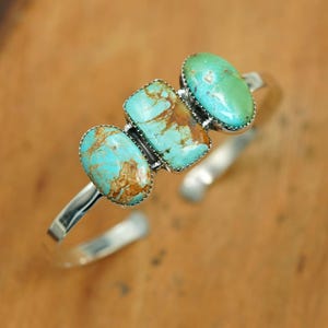 May include: A silver cuff bracelet with three turquoise stones. The stones are oval and rectangular, with blue and brown patterns. The open-ended bracelet is displayed on a wooden surface.