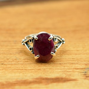 May include: A silver ring featuring an oval, deep red gemstone. The ring's band has an ornate, swirling design on either side of the gemstone. The ring is set against a warm, wooden surface.