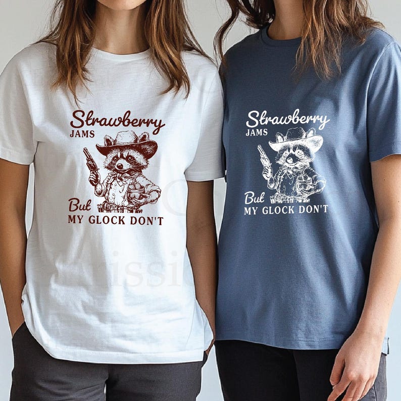 May include: Two white t-shirts with a blue t-shirt in the background. The white t-shirts feature a cartoon raccoon wearing a cowboy hat and holding a gun. The text on the shirts reads "Strawberry Jams But My Glock Don't".