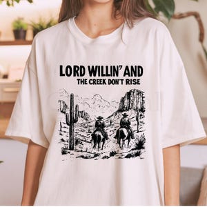 May include: White t-shirt with a black graphic print of two cowboys riding horses in a desert landscape. The text "Lord Willin' and the Creek Don't Rise" is printed above the graphic.