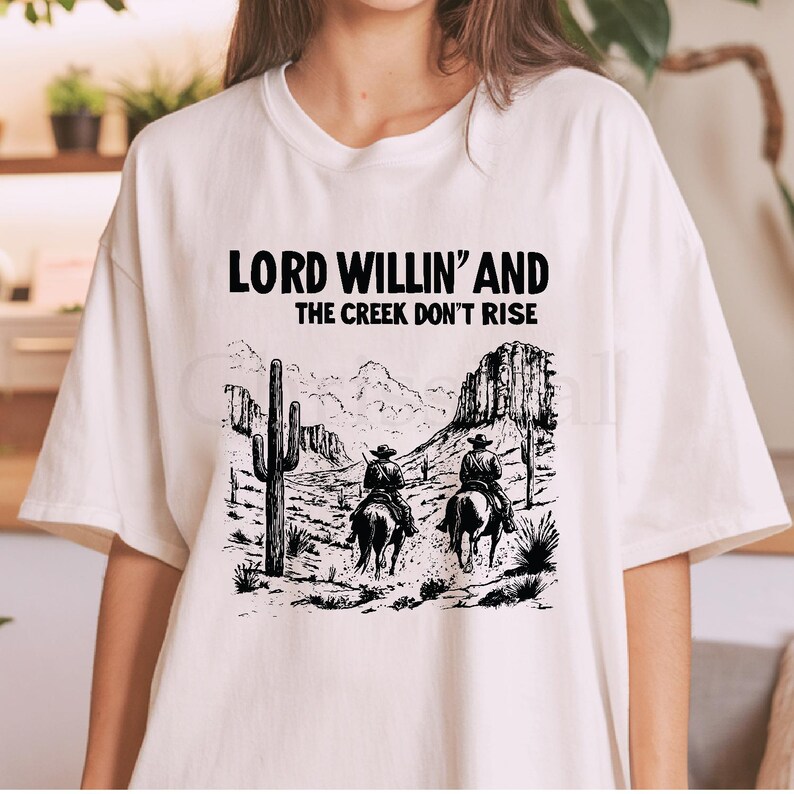 May include: White t-shirt with a black graphic print of two cowboys riding horses in a desert landscape. The text "Lord Willin' and the Creek Don't Rise" is printed above the graphic.